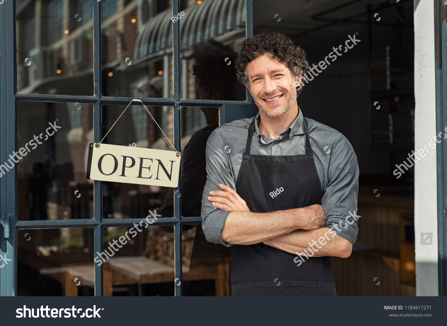 Portrait of small business owner smiling and standing with crossed arms outside the cafe. Portrait of handsome mature waiter standing in entrance of coffee shop.  #1184817271
