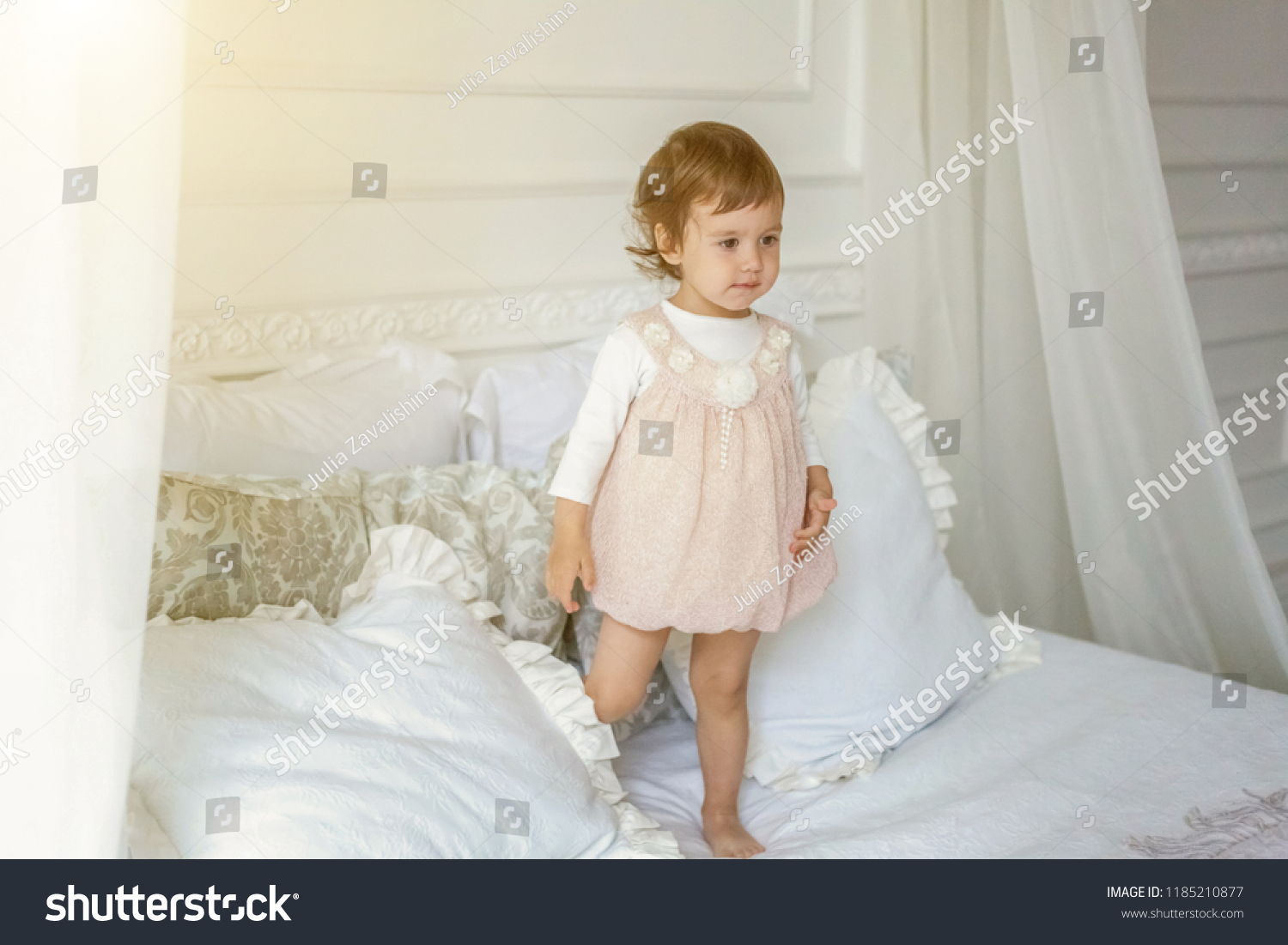 Funny and cute brunette little smiling girl playing jumping on bed in light bedroom. White interior with big bed. Childhood, preschool, youth, relax concept #1185210877