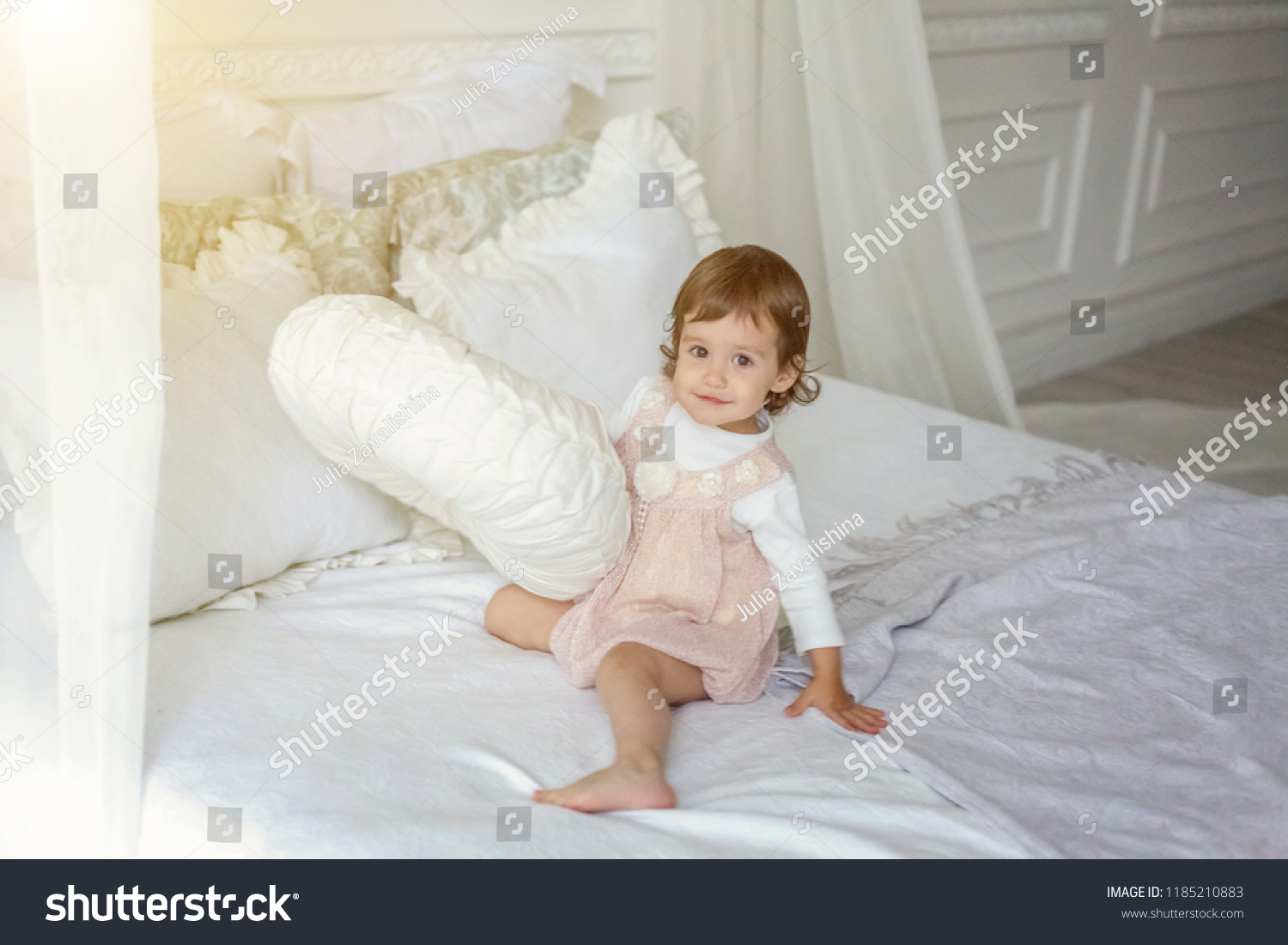 Funny and cute brunette little smiling girl playing jumping on bed in light bedroom. White interior with big bed. Childhood, preschool, youth, relax concept #1185210883