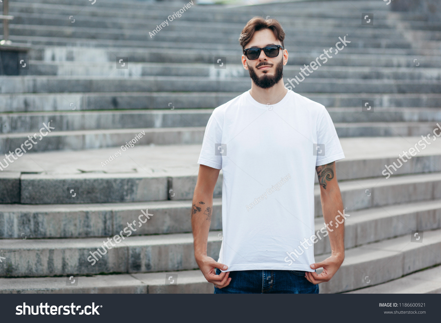 A young stylish man with a beard in a white T-shirt and glasses. Street photo #1186600921