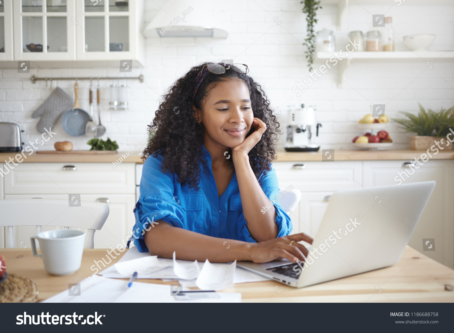 Indoor shot of happy young Afro American woman using portable computer and smiling, enjoying modern technology while paying bills for rent, gas and electricity online, sitting at kitchen table #1186688758