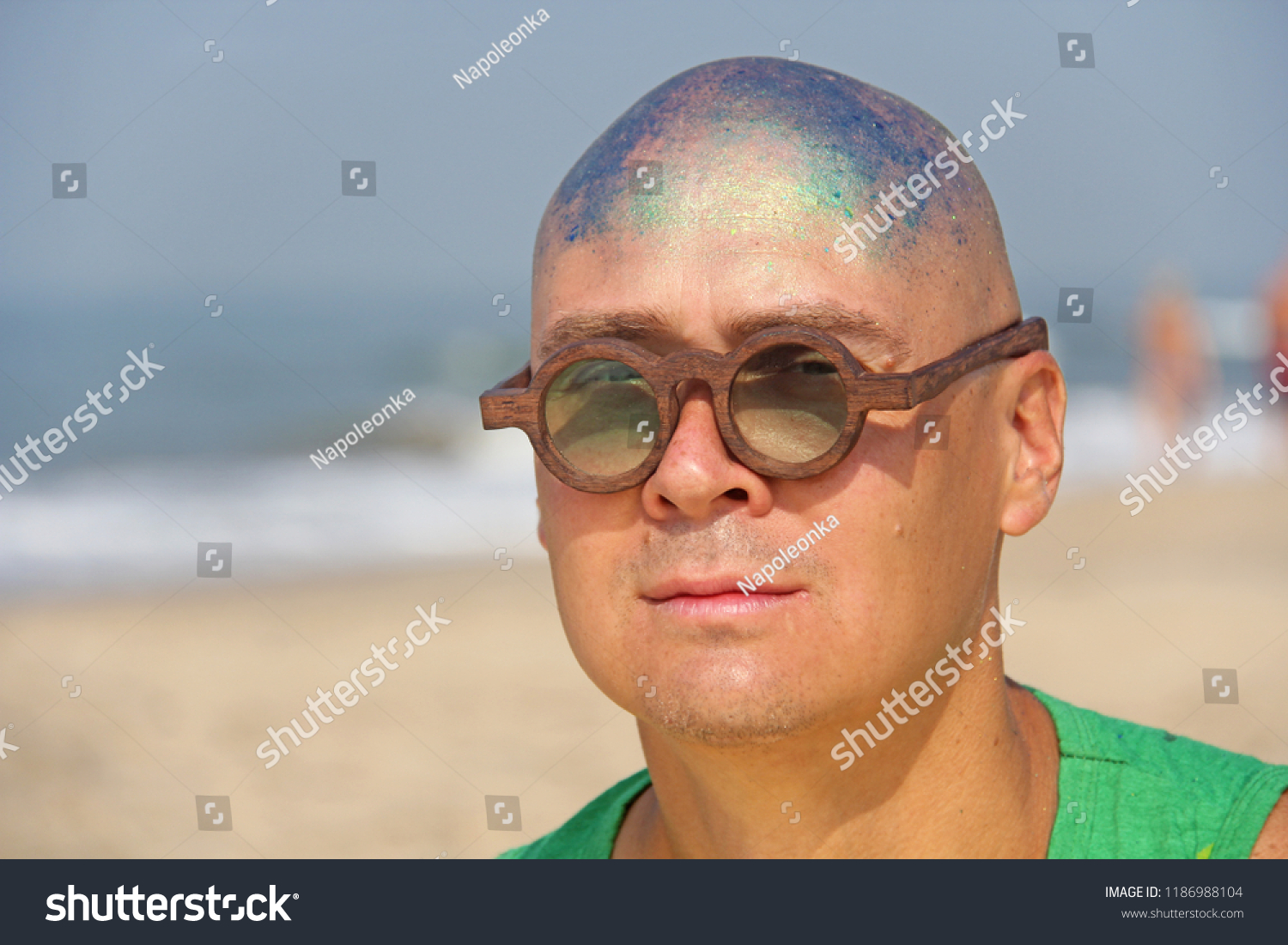 A bald and unusual young man, a freak, with a shiny bald head and round wooden glasses on the background of the beach and the sea. Humor and eccentricity. Unusual appearance. Humorist. unexpected. #1186988104