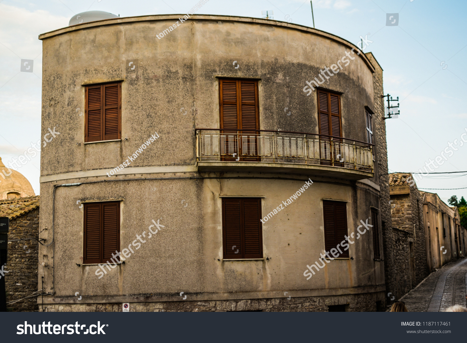 Interesting, strange, rounded house building in Erice, Sicily, Italy. Weird type of architecture, with the balcony on the 1st floor.  In the background narrow, Italian city street. Ancient style.  #1187117461