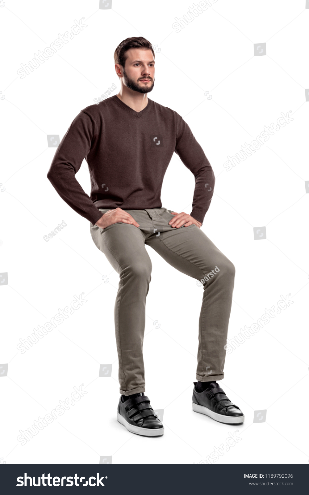 An isolated bearded man in casual wear sits on a white background with hands on his thighs. Sitting and waiting. Meditating. Neutral expression. #1189792096