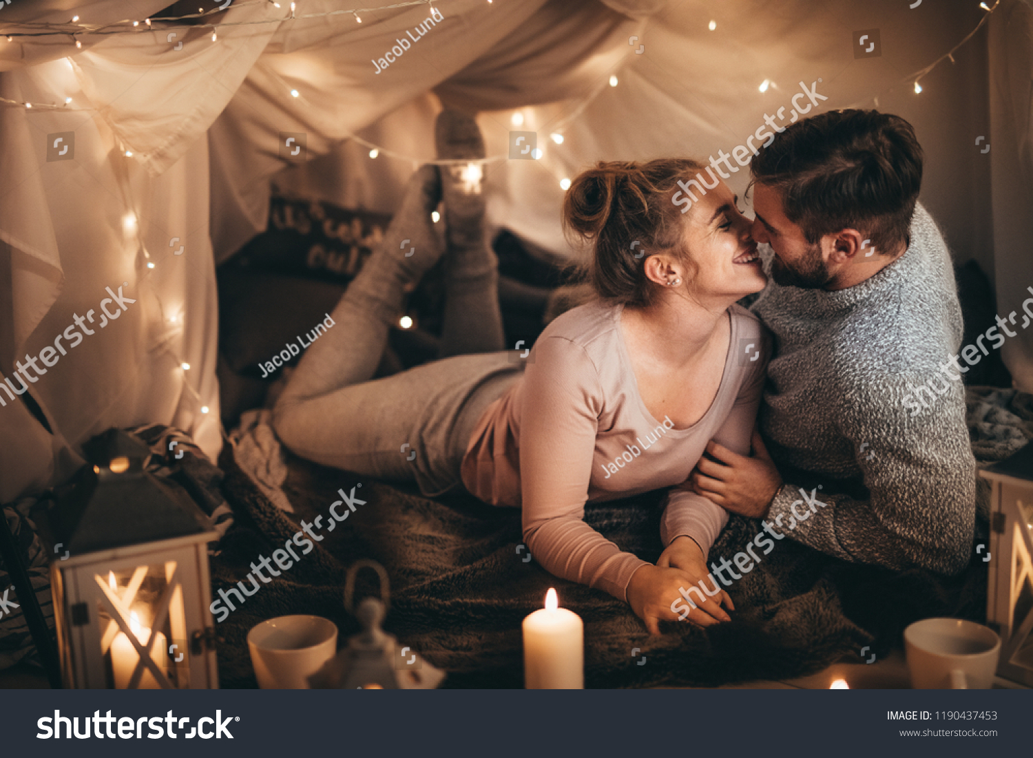 Couple together on bed in a room decorated with candle lights and tiny serial light bulbs. Smiling woman spending happy time with her husband in bedroom. #1190437453