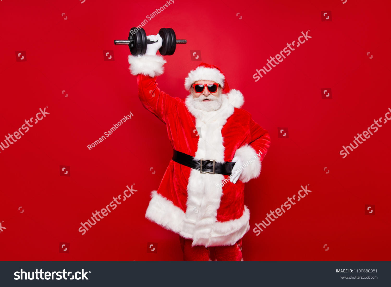 Cheerful funny trendy stylish fashionable strong sporty virile muscular Santa in eyeglasses gloves fur white red winter coat black belt lifting one big dumbbell striving isolated on red background #1190680081