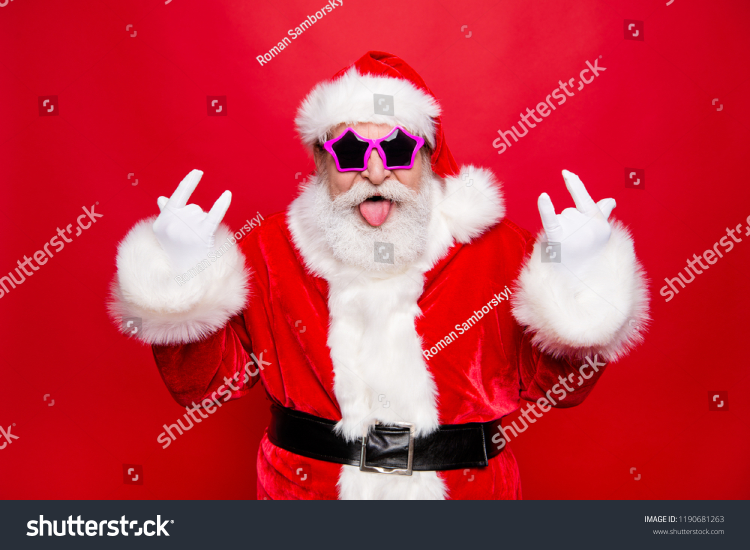 Time to dance on heavy metal disco music! Mature grandfather Santa in costume glasses with playful mood fooling around show tongue out make rock n roll sign isolated december noel red background #1190681263