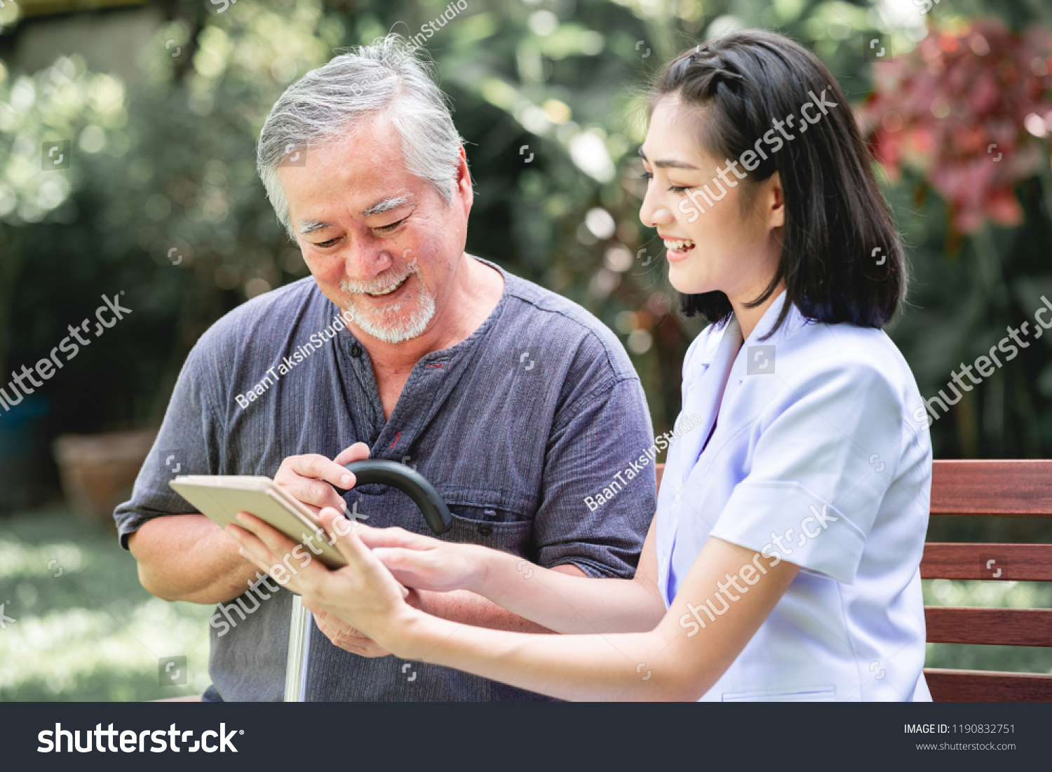 Nurse with patient sitting on bench together looking at tablet. Asian old man and young woman sitting together talking. Relax mood. #1190832751