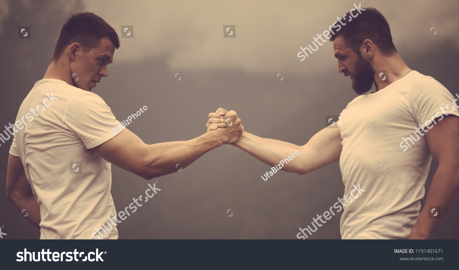 Strong male bodybuilders in white t-shirts greeting each other clasping hands together in outdoor workout over foggy mountain landscape. Rivalry, challenge, strength comparison concept #1191401671