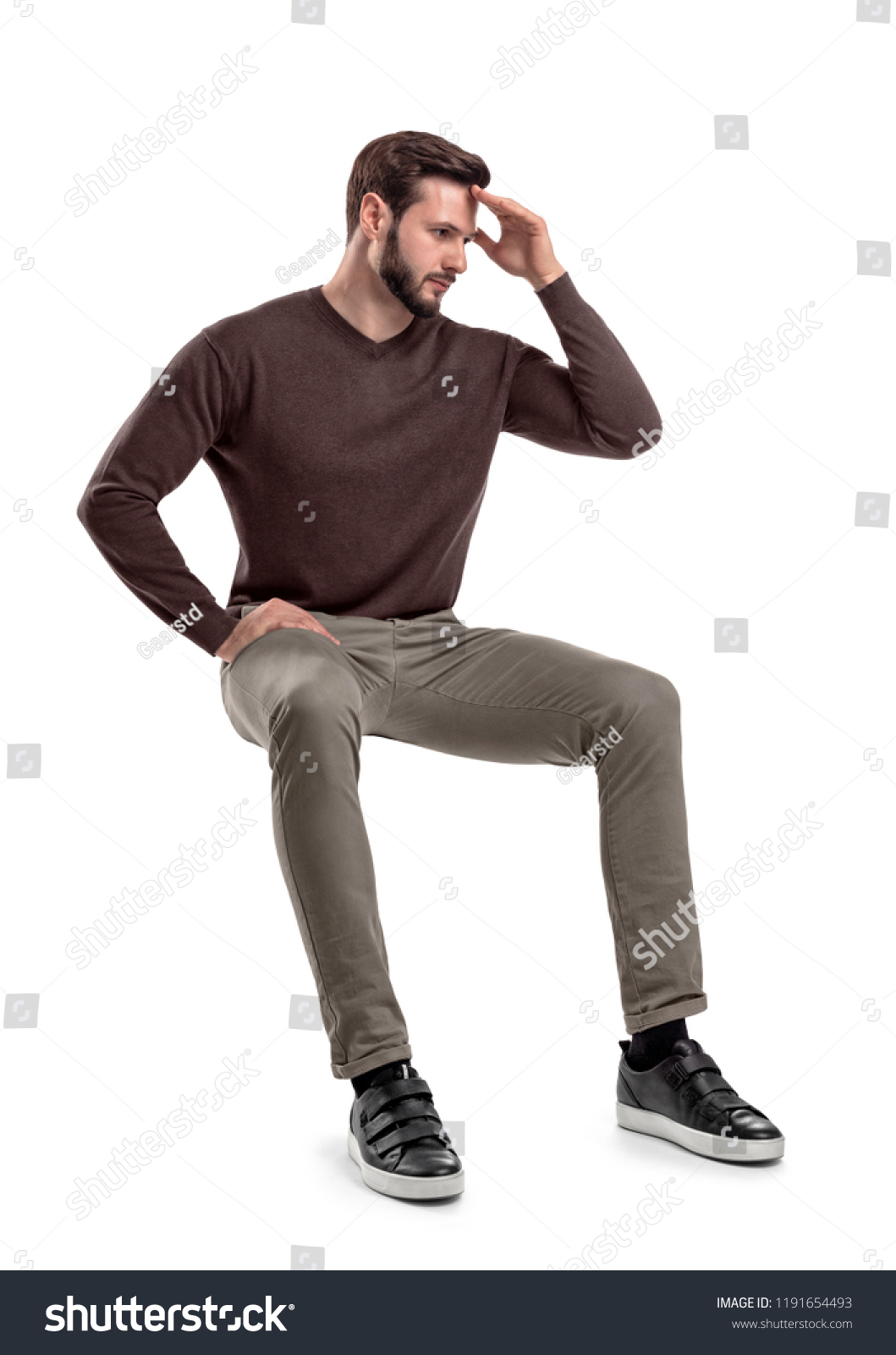 An isolated bearded man in casual wear sits on a white background with one hand touching his forehead. Thinking about troubles. Unresolved problems. Headache. #1191654493