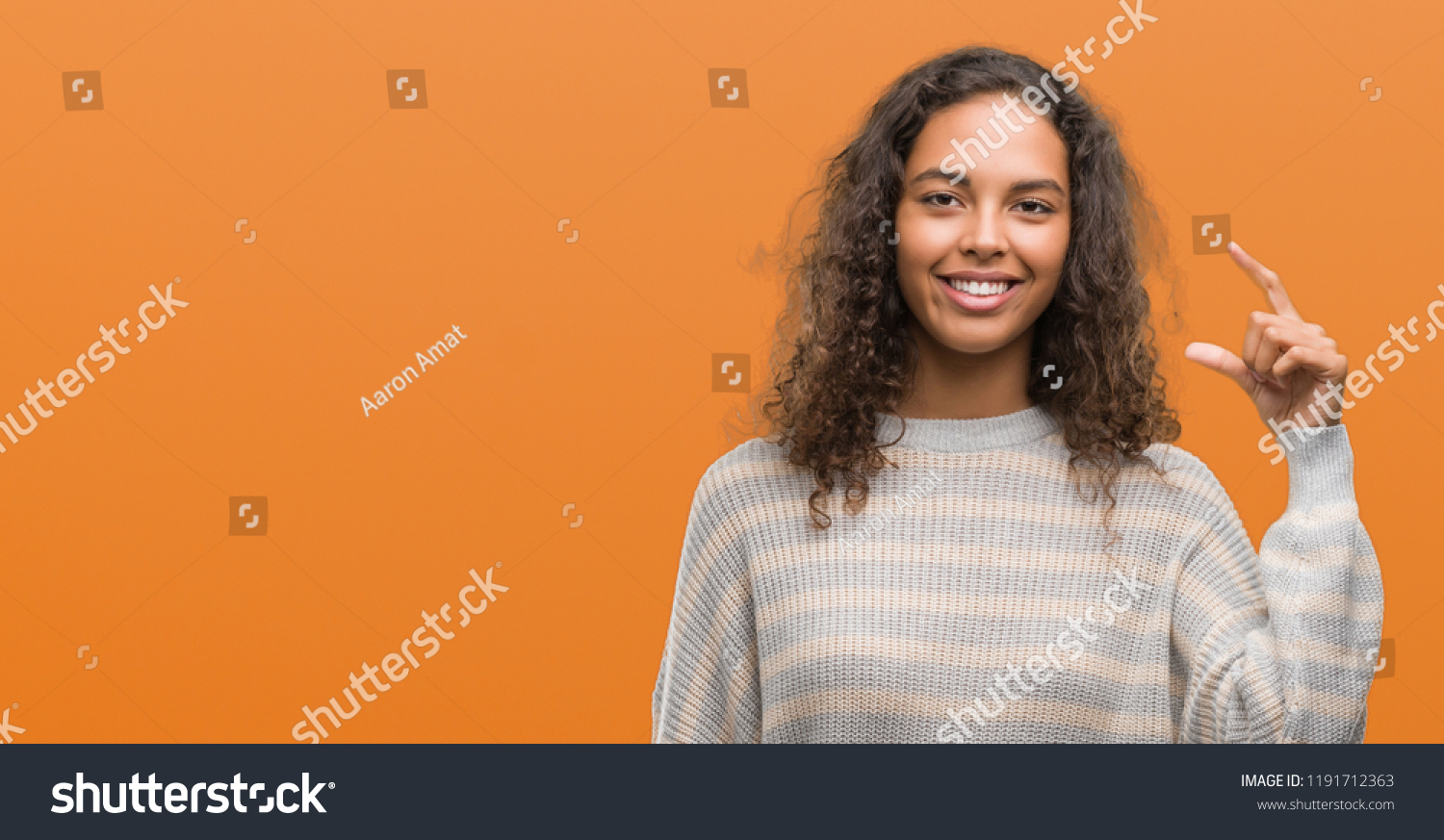 Beautiful young hispanic woman wearing stripes sweater smiling and confident gesturing with hand doing size sign with fingers while looking and the camera. Measure concept. #1191712363