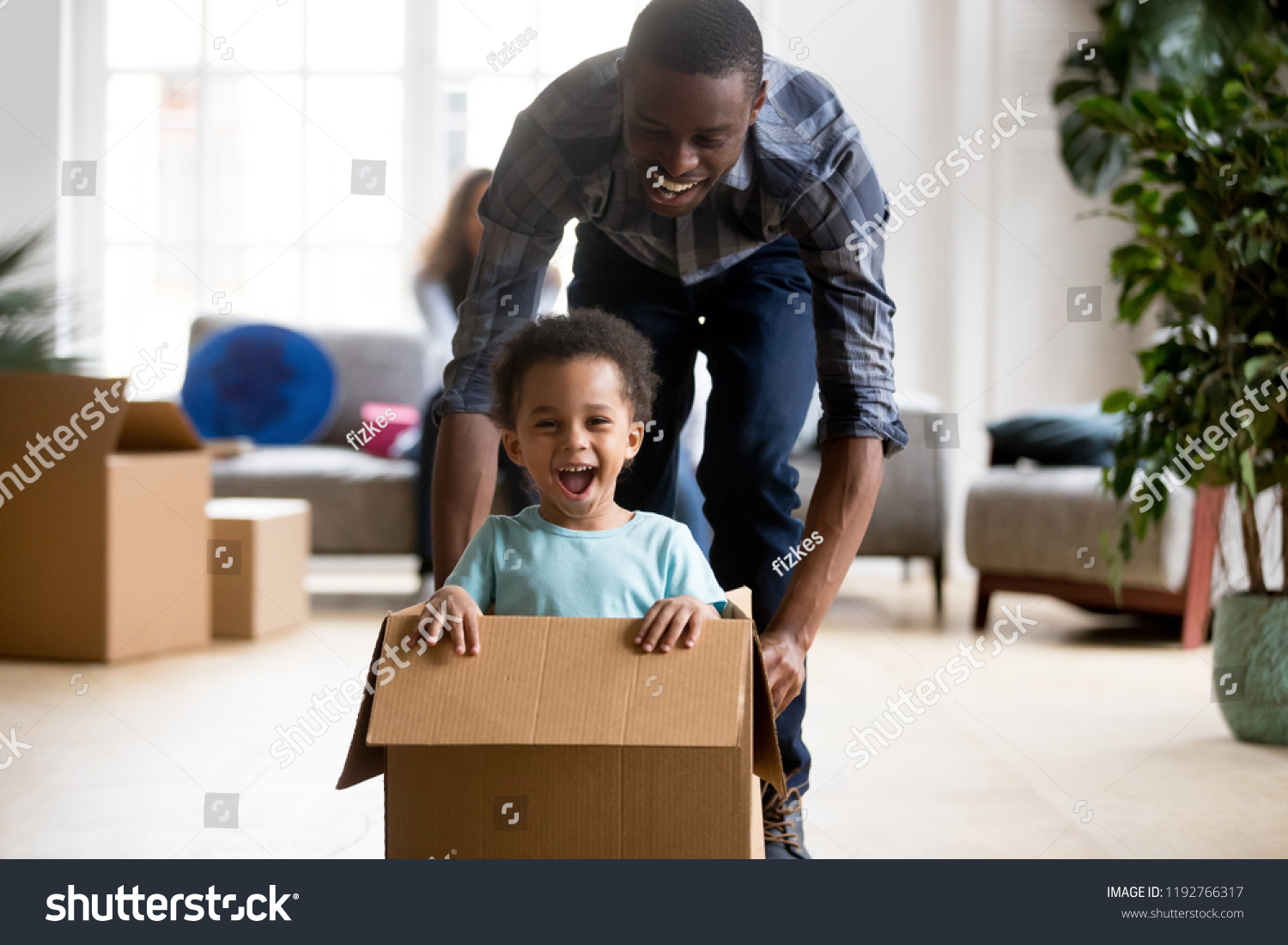 Black family in living room have a fun spend time at new home. African adorable playful laughing boy sitting at cardboard box, father rolling him playing together. New property and relocation concept #1192766317