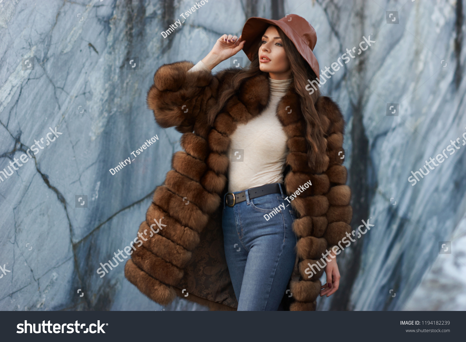 Young gorgeous woman with long wavy brunette hair in brown fur coat standing and posing against gray marble walls at stone quarry. Rich expensive woman #1194182239