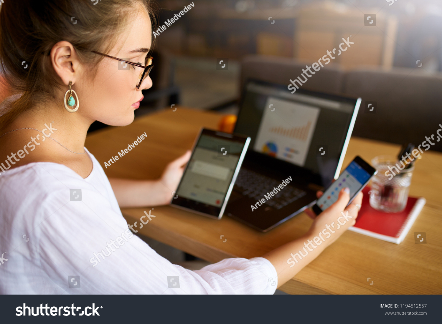 Mixed race woman in glasses working with multiple electronic internet devices. Freelancer businesswoman has tablet and cellphone in hands and laptop on table with charts on screen. Multitasking theme #1194512557