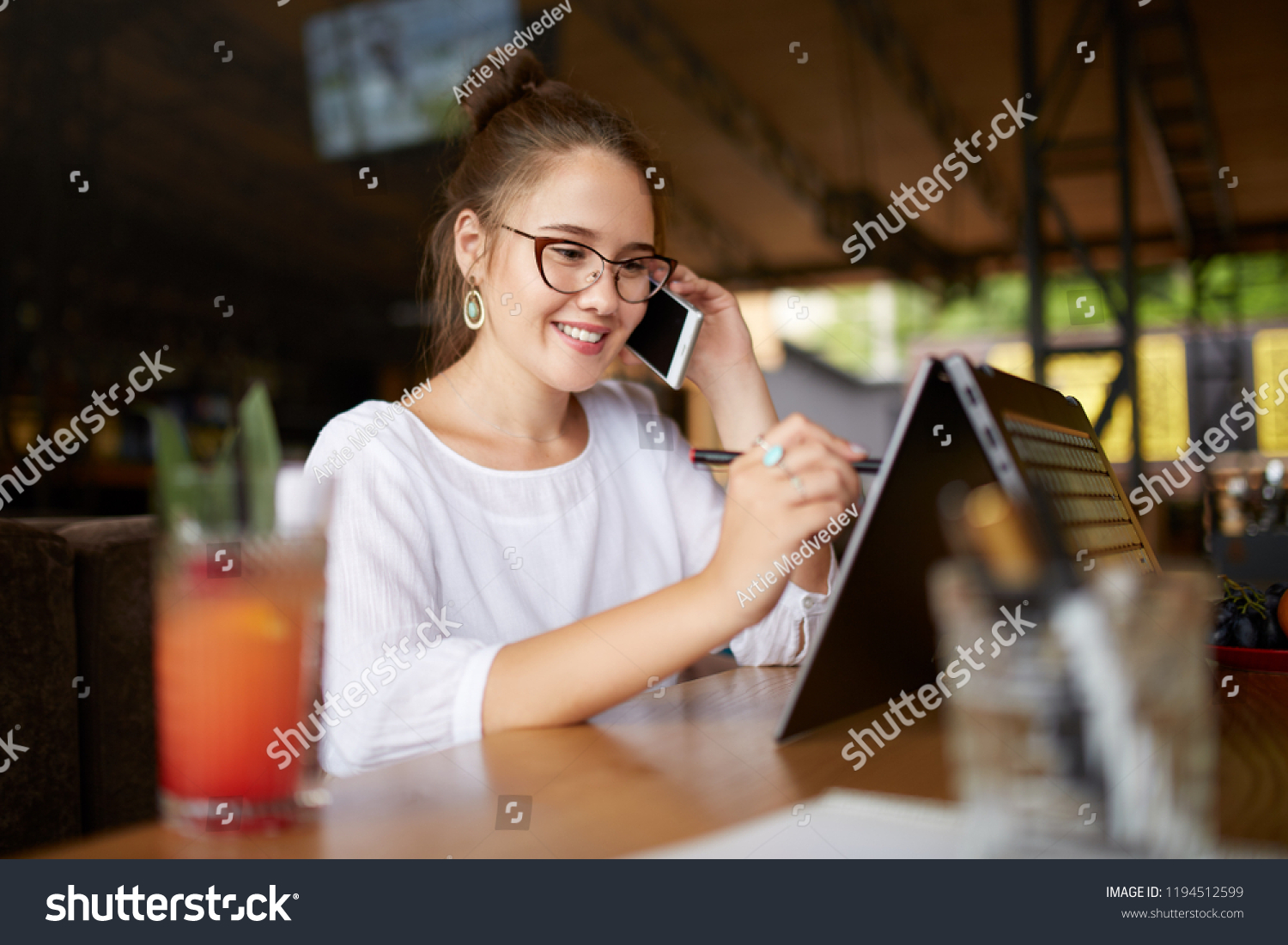 Mixed race freelancer working with convertible laptop and talking on cellphone with client in cafe. Asian caucasian businesswoman conducts negotiations via phone call. Multitasking business concept. #1194512599