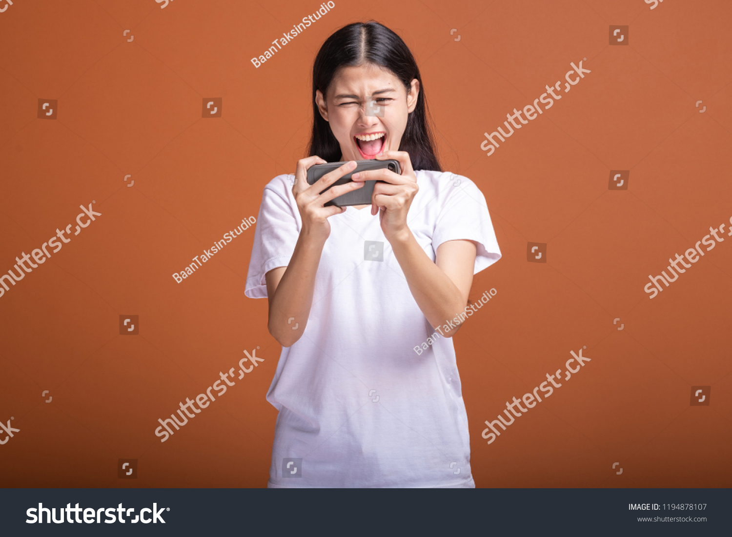Woman playing mobile game isolated over orange background. Young asian woman in white t-shirt, upset mood. Young hipster lifestyle concept. #1194878107