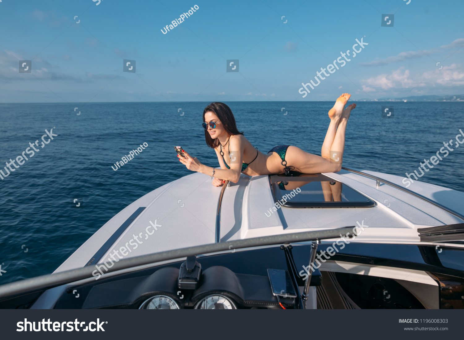 Vacation and technology. Pretty young woman in swimsuit chatting with a friend while taking sun bathes on a bow of a luxury sailboat. #1196008303