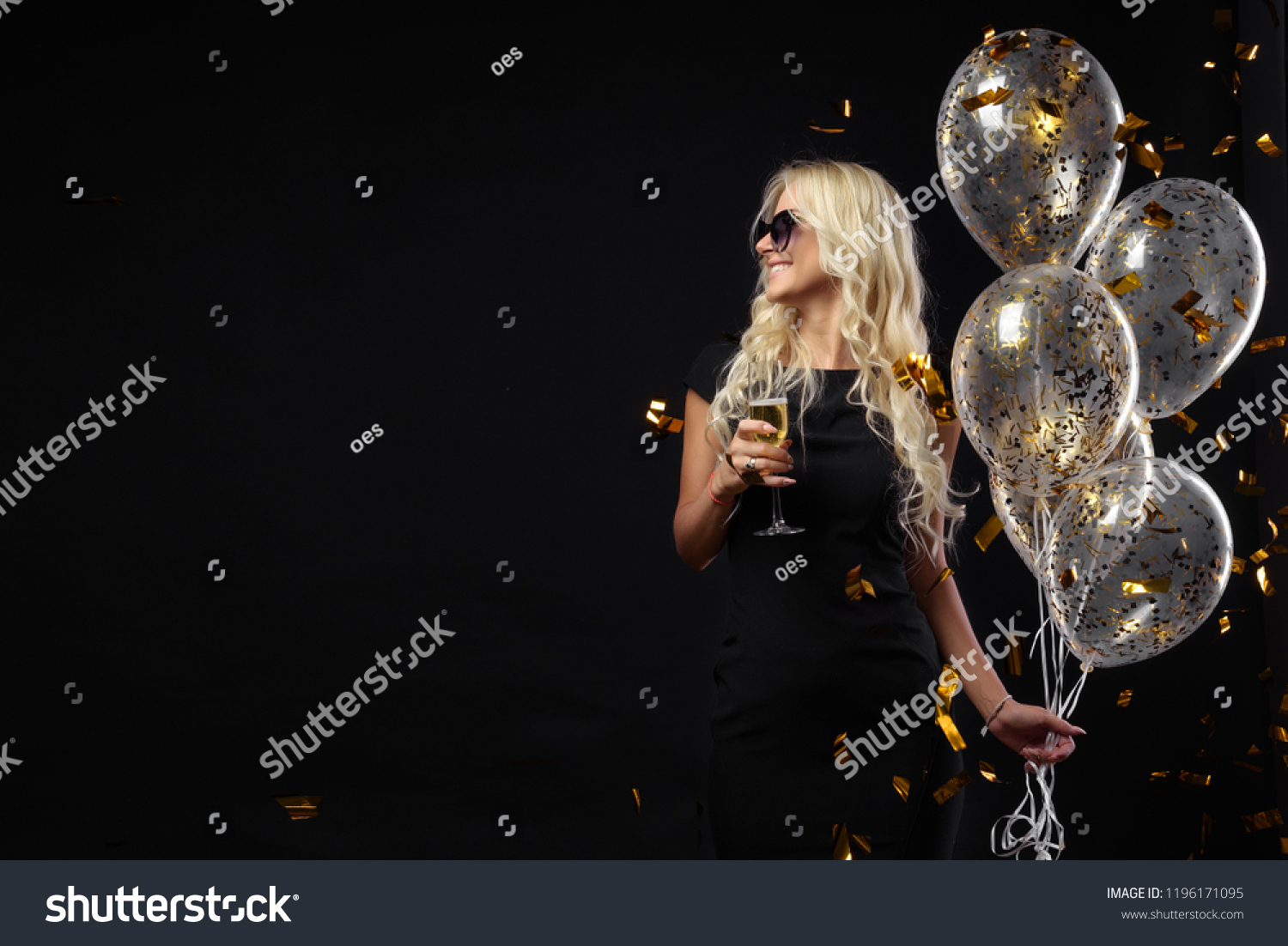 Brightfull expressions of happy emotions of  amazing blonde girl celebrating party on black background. Luxury black dresses, smiling, a glass of champagne, golden tinsels,  balloons, long curly hair #1196171095
