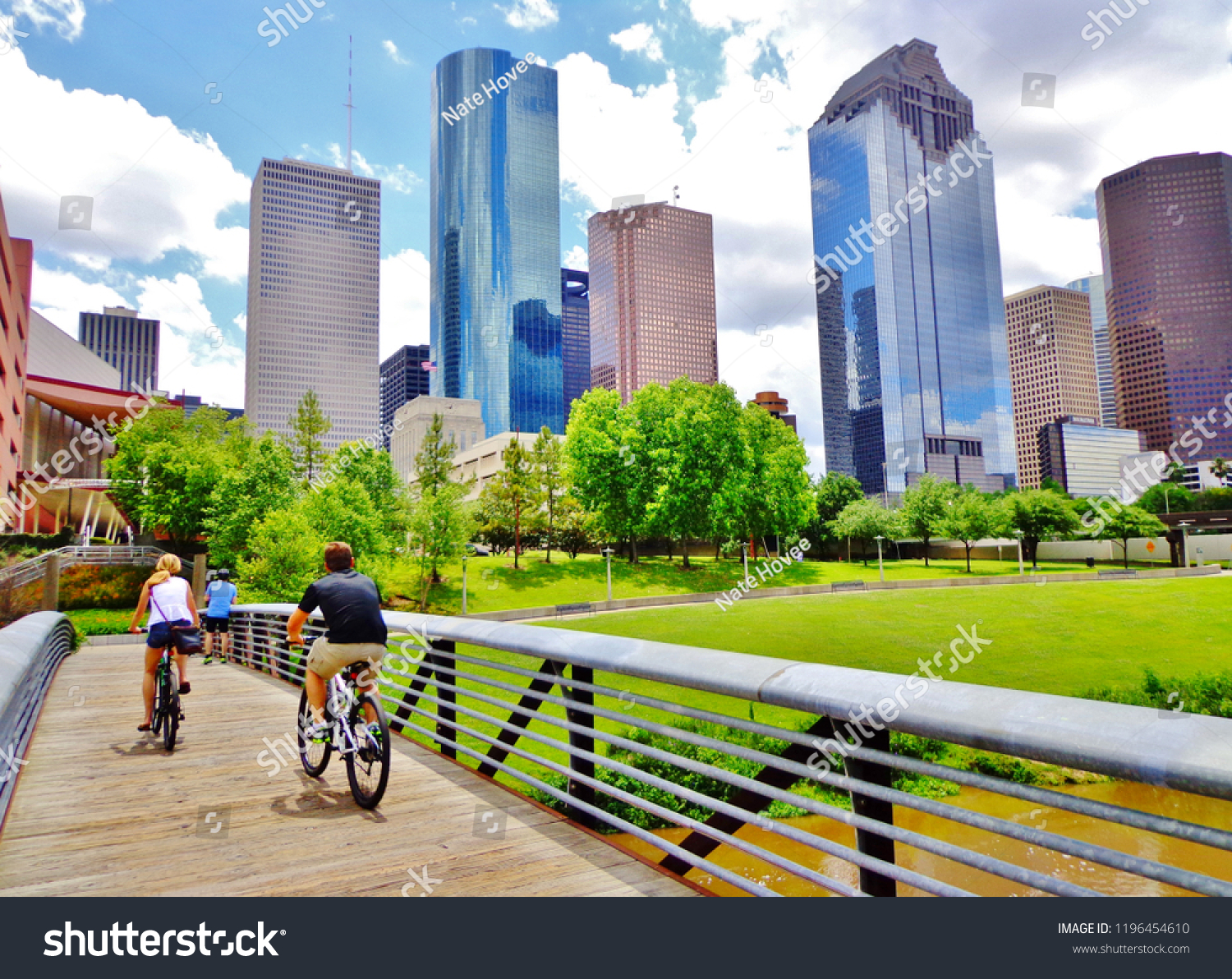 Bicyclists cross wooden bridge in Buffalo Bayou Park, with a beautiful view of downtown Houston (skyline / skyscrapers) in background on a summer day - Houston, Texas, USA  #1196454610