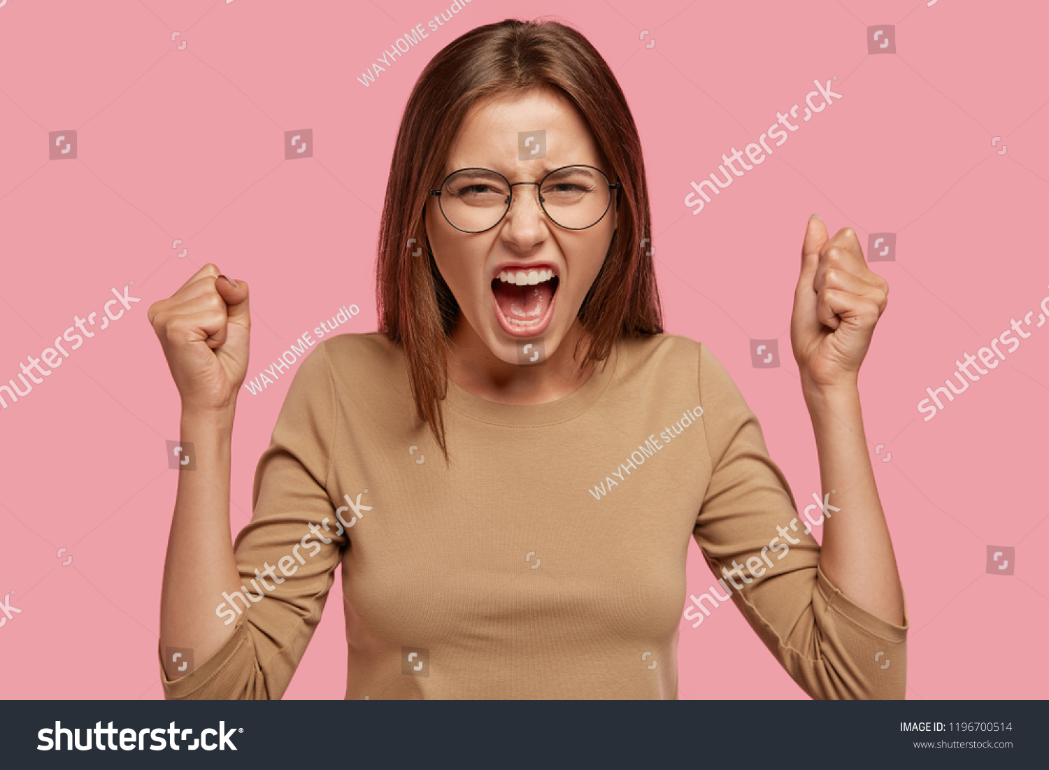 Photo of annoyed ourtraged housewife has quarrel with neighbour, raises hands clenched in fists, screams with anger, isolated over pink background, wears round transparent glasses. Negative feeling #1196700514