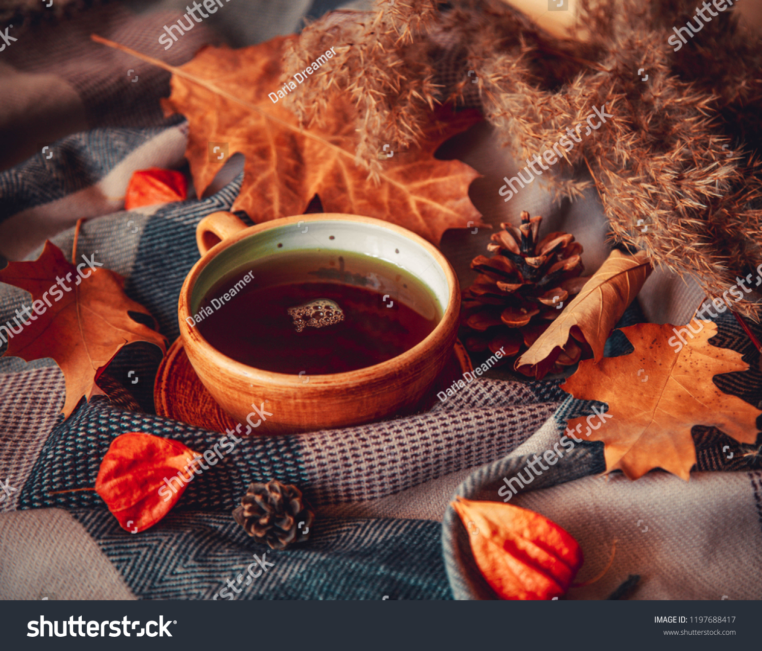 Autumn concept image with hot mug of tea, leaves, pinecones, physalis and warm scarf. Warm drink in cold weather. Home decoration.  #1197688417