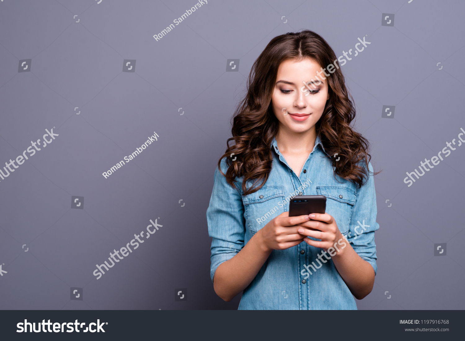 Content calm trendy nice cute adorable lovely attractive brunette caucasian girl with wavy hair in casual denim shirt, playing game in phone, isolated over grey background #1197916768