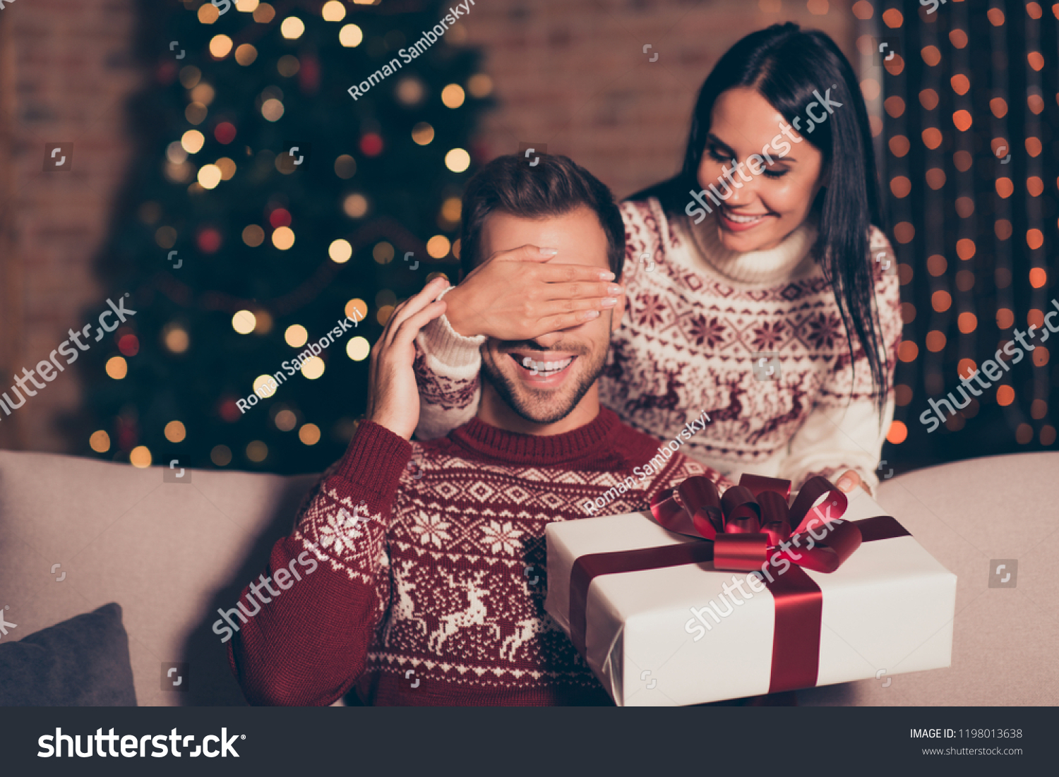 Hide and seek concept. Good-looking, attractive brunette lady in ornament sweater hold big package with bow, close eyes her man, who sit in cozy living room couch with lights garland decorations #1198013638