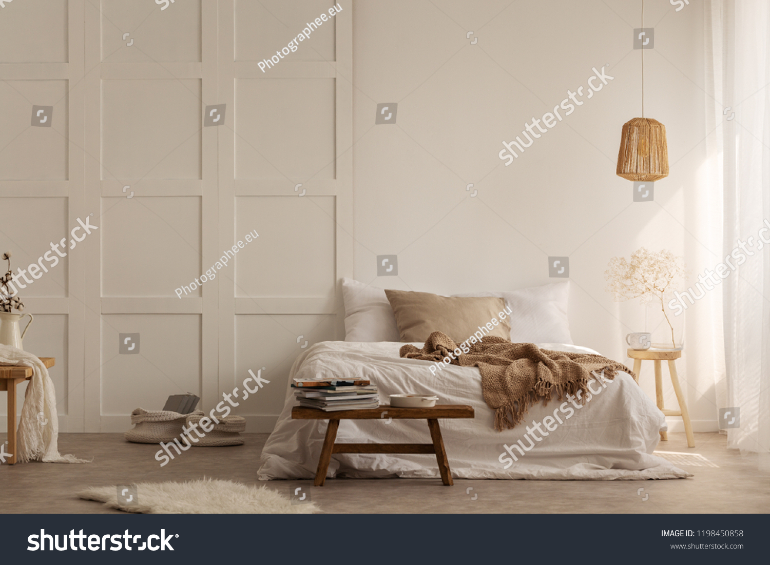 Natural blanket on white bed in simple bedroom interior with fur next to wooden stool. Real photo #1198450858