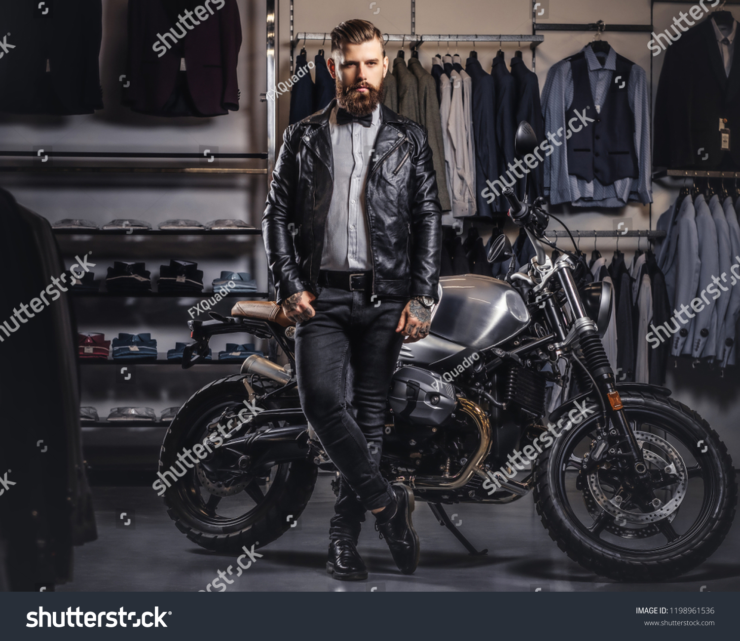 Stylish tattooed bearded man with dressed in black leather jacket and bow tie posing near retro sports motorbike at men's clothing store. #1198961536