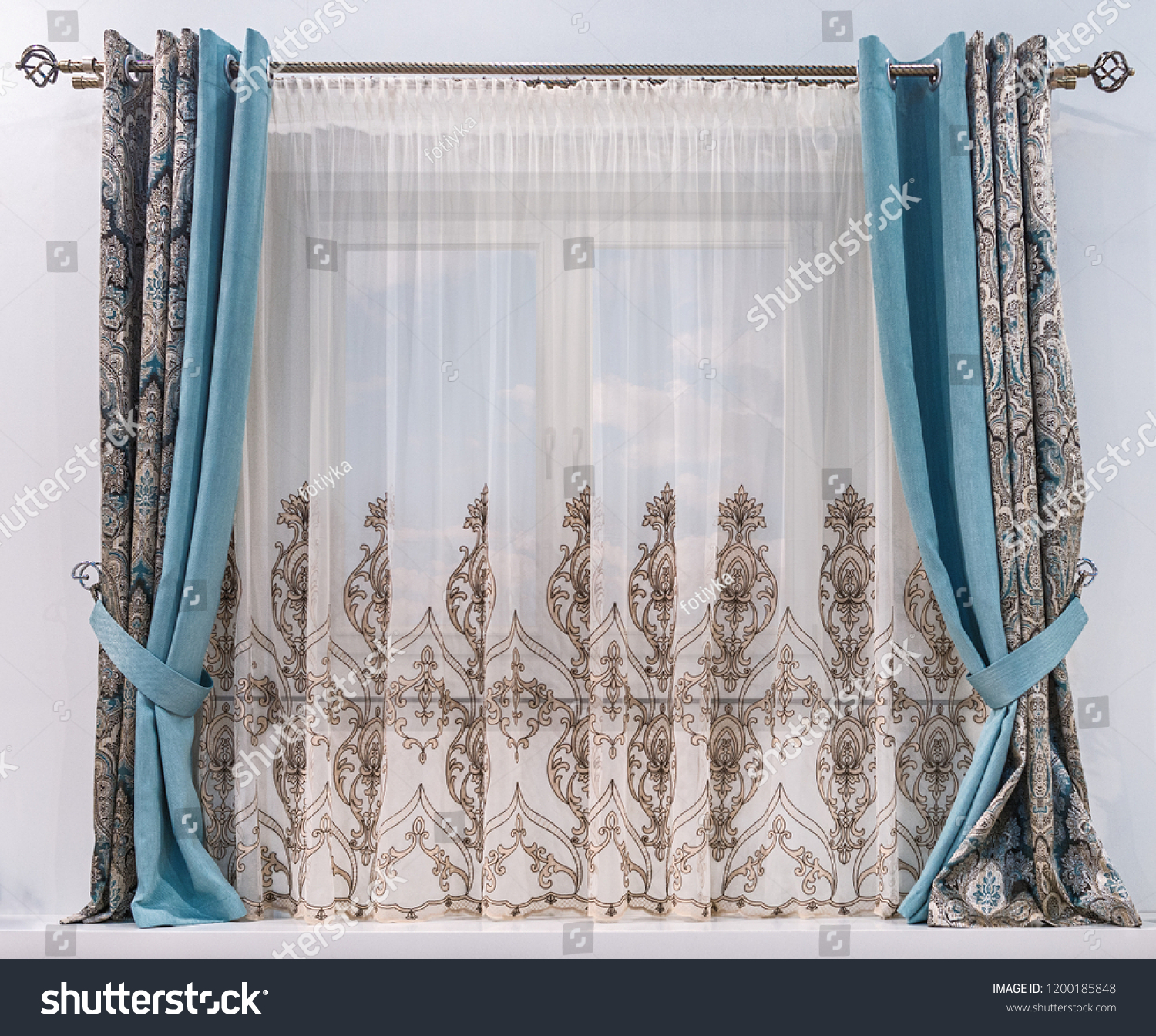Modern design of a small window. Combined curtain with eyelets, monochromatic turquoise fabric, tapestry with an ornament and a tulle with patterns hang on a round cornice. #1200185848