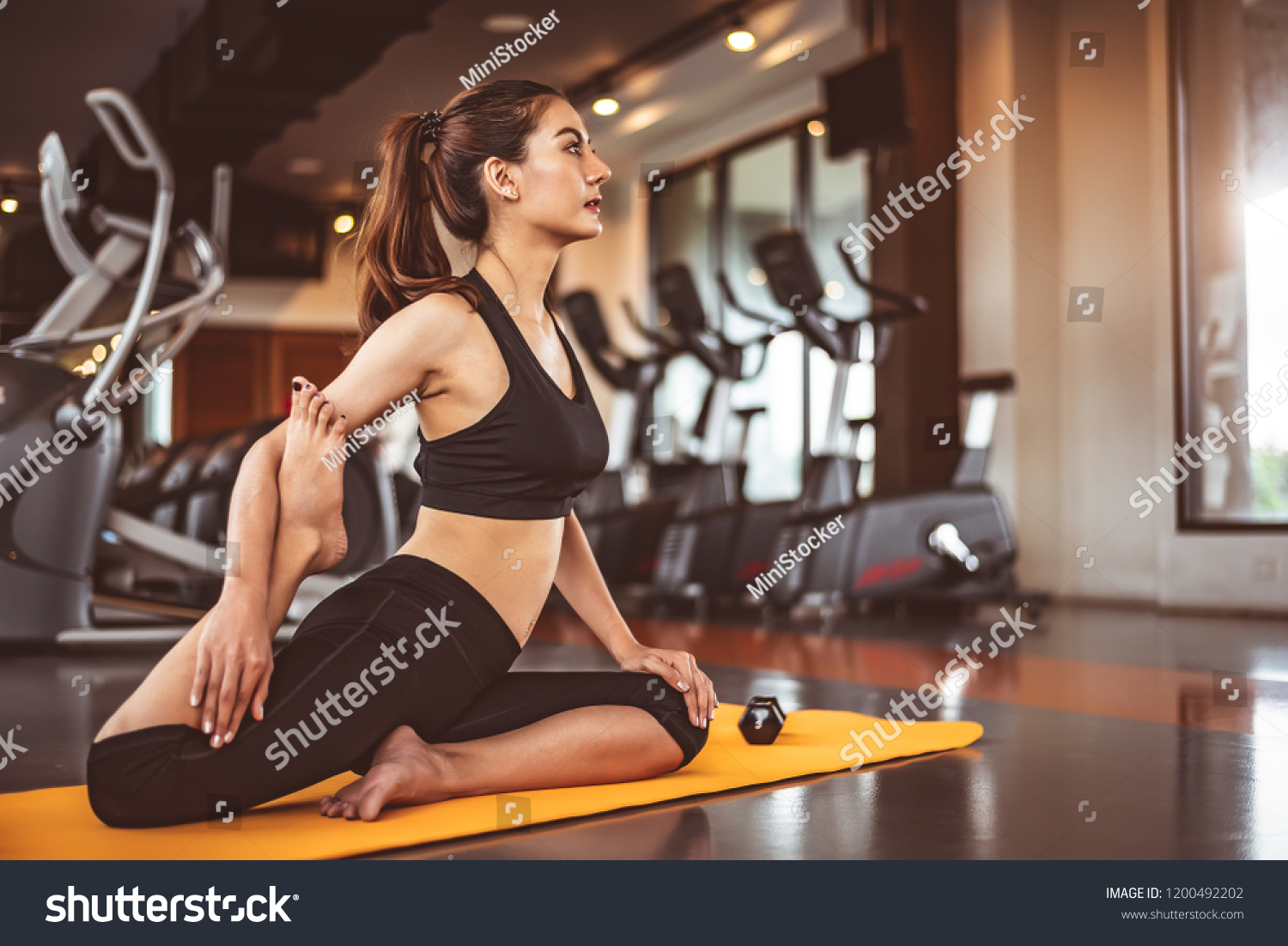 Woman doing bending legs yoga in fitness workouts training gym center background. Lifestyle woman sitting with sport dumbbell equipment and exercise treadmill background. Yoga girl in black sports bra #1200492202