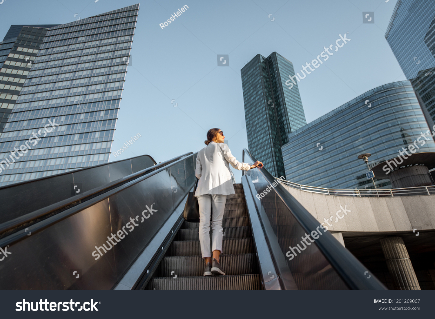 Stylish businesswoman in white suit going up on the escalator at the business centre outdoors with skyscrapers on the background in Paris #1201269067