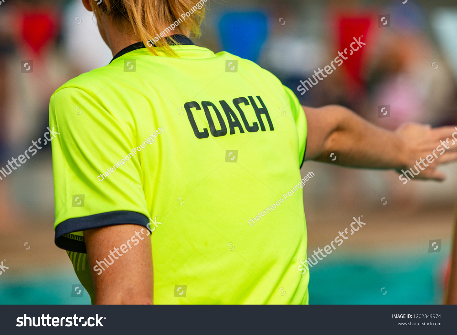 Back view of female swim coach, wearing COACH shirt, giving instruction to her swimmers #1202849974