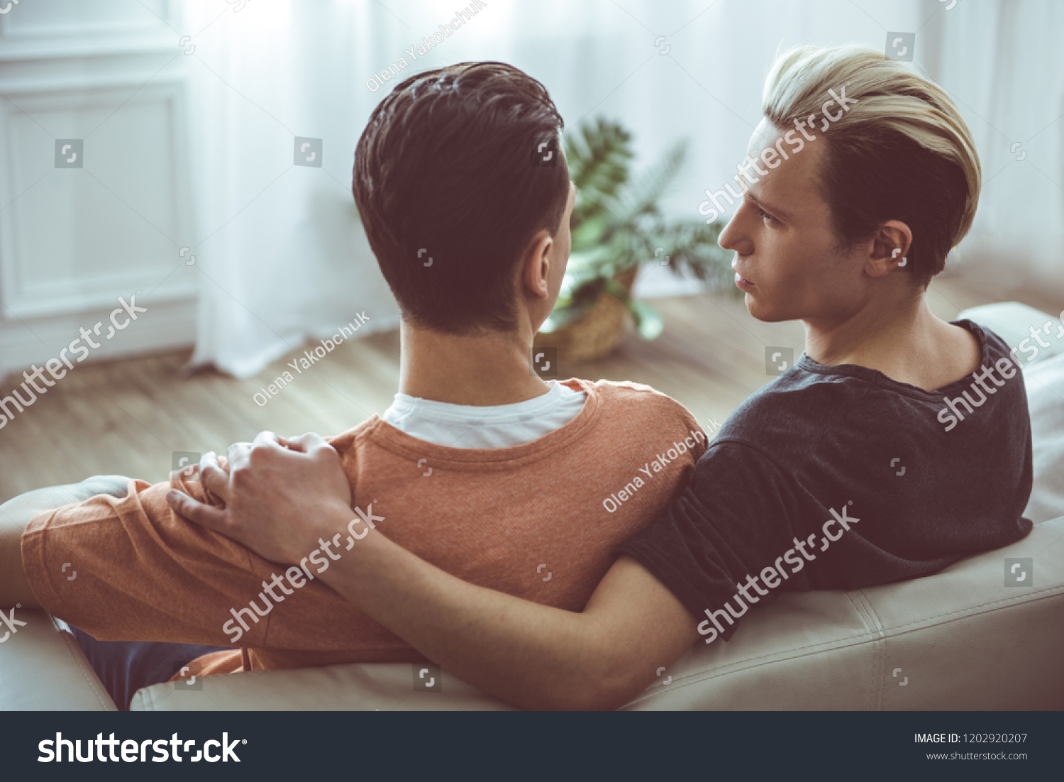 Back view portrait of young man with dyed hair hugging boyfriend and gazing at him. They are resting on sofa at home #1202920207