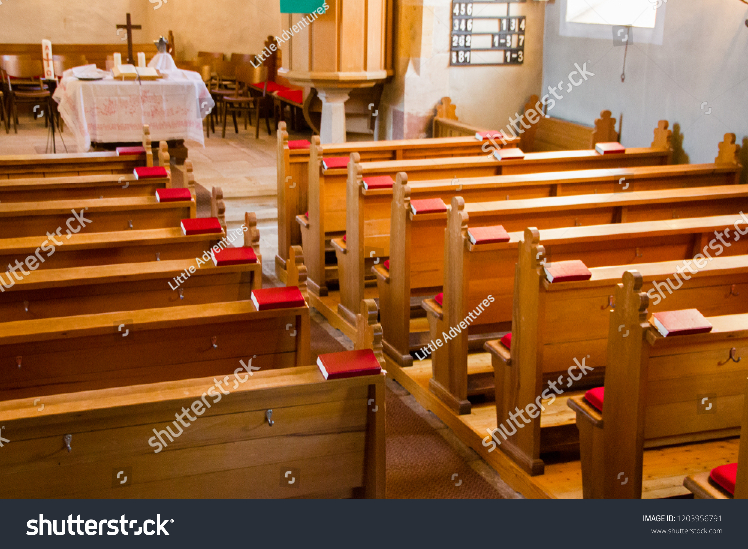 Empty wooden church benches of a Christian church with bible or choir books with cross . Blurry alter in the background. Selective focus. Concept of church service or empty churches. #1203956791
