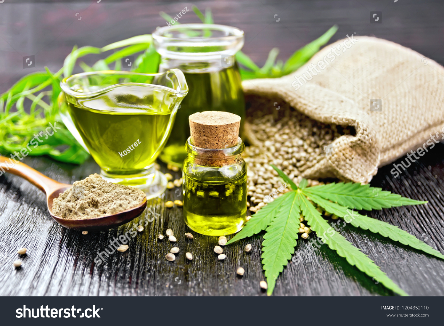 Hemp oil in two glass jars and sauceboat with grain in the bag, leaves and stalks of cannabis, a spoon with flour on the background of wooden boards #1204352110