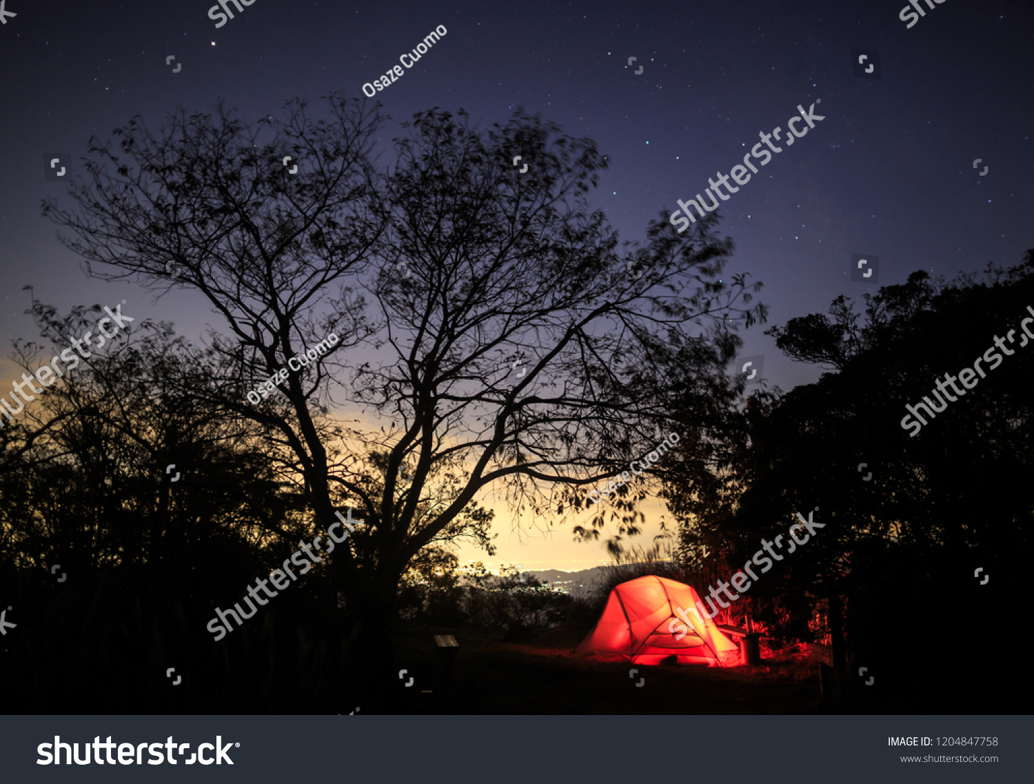 Tent glows red against distant glow from city and starry night sky #1204847758