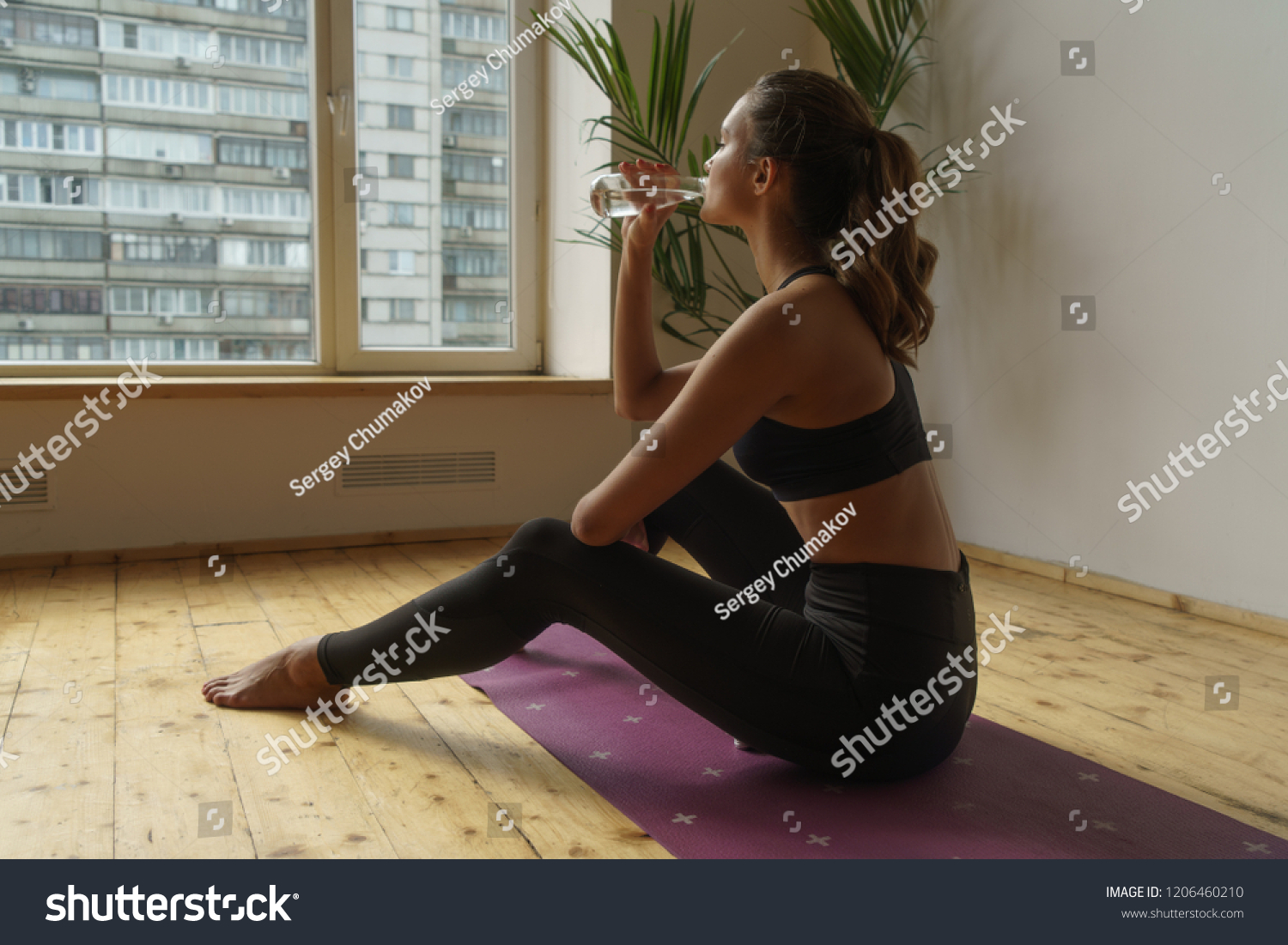 young sportive woman sitting on yoga carpet and drinking clear water at home #1206460210