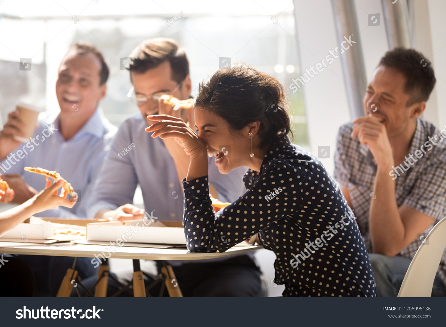 Indian woman laughing at funny joke eating pizza with diverse coworkers in office, friendly work team enjoying positive emotions and lunch together, happy colleagues staff group having fun at break #1206996136