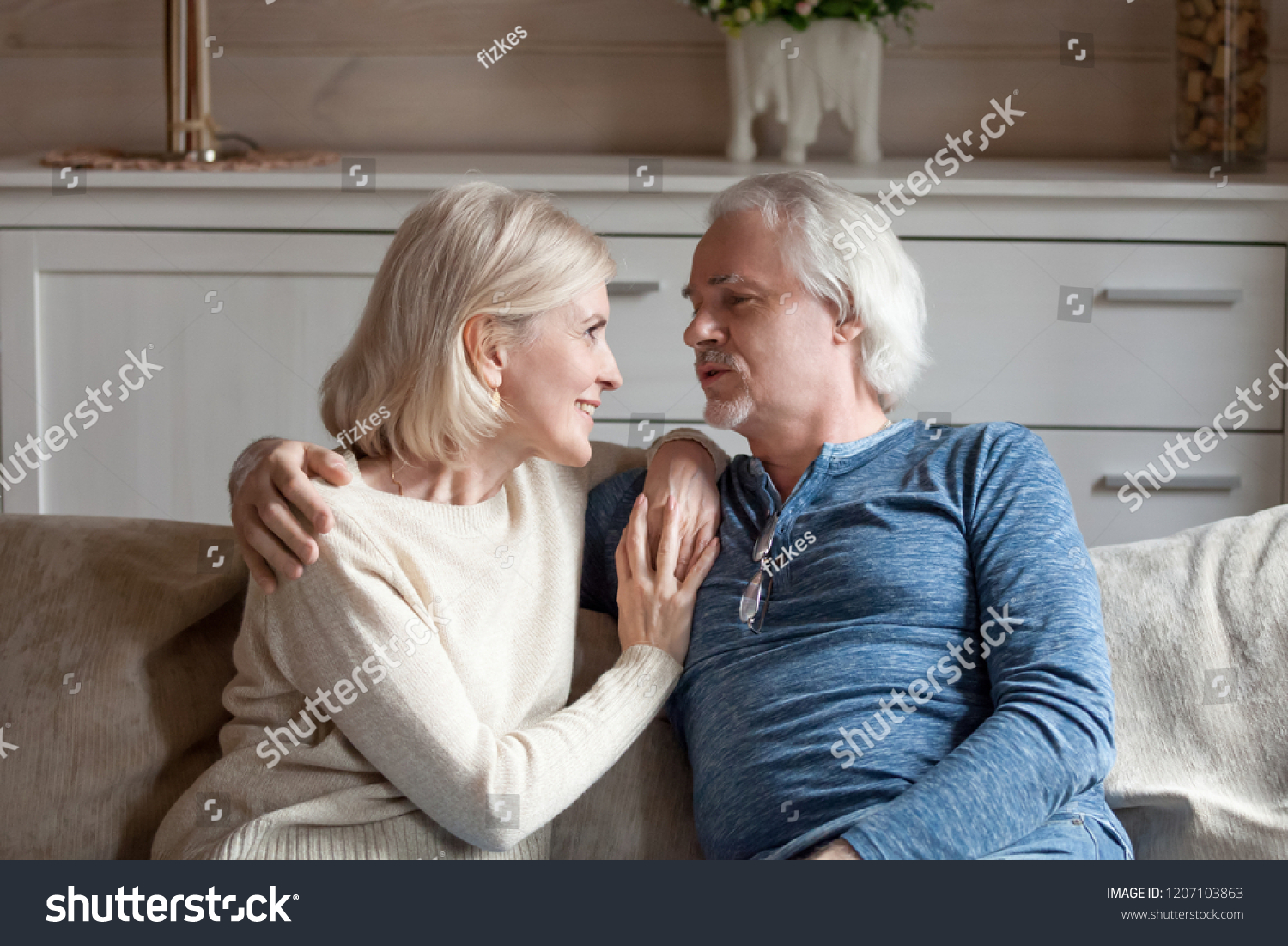 Happy senior husband and wife look at each other talking relaxing on couch, smiling aged couple cuddle and embrace on sofa at home, romantic elderly man and woman enjoy rest having conversation #1207103863