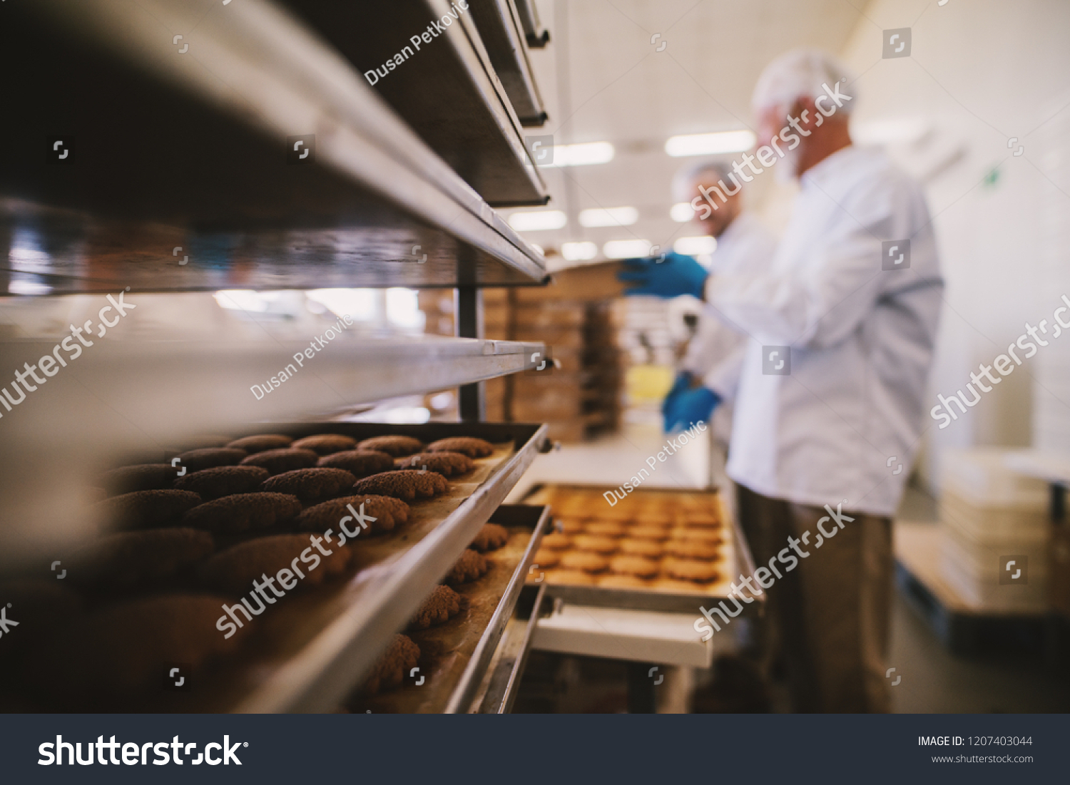 Close up of tray full of fresh baked cookies in food factory. Blurred picture of two male employees in sterile clothes in background. #1207403044