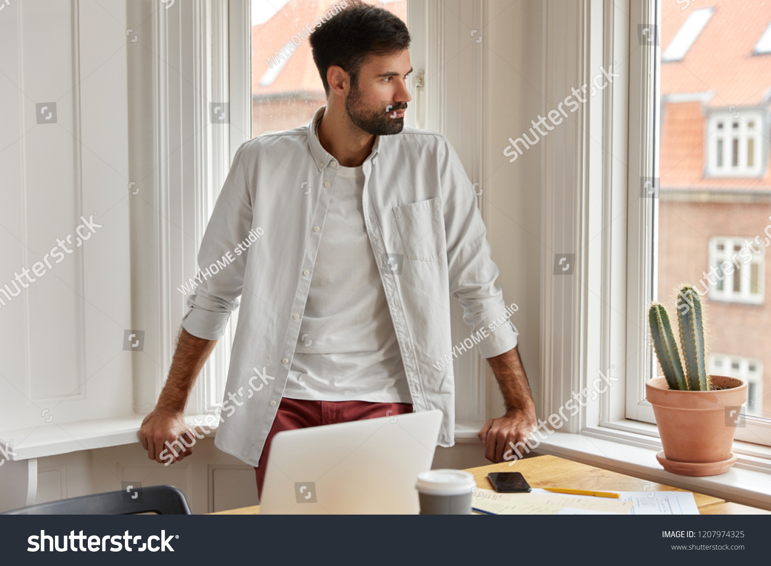 Shot of male employer works from home, stands near big window and desktop with laptop computer, watches conference video, studies documentation, has thoughtful look aside. Busy financier indoor #1207974325