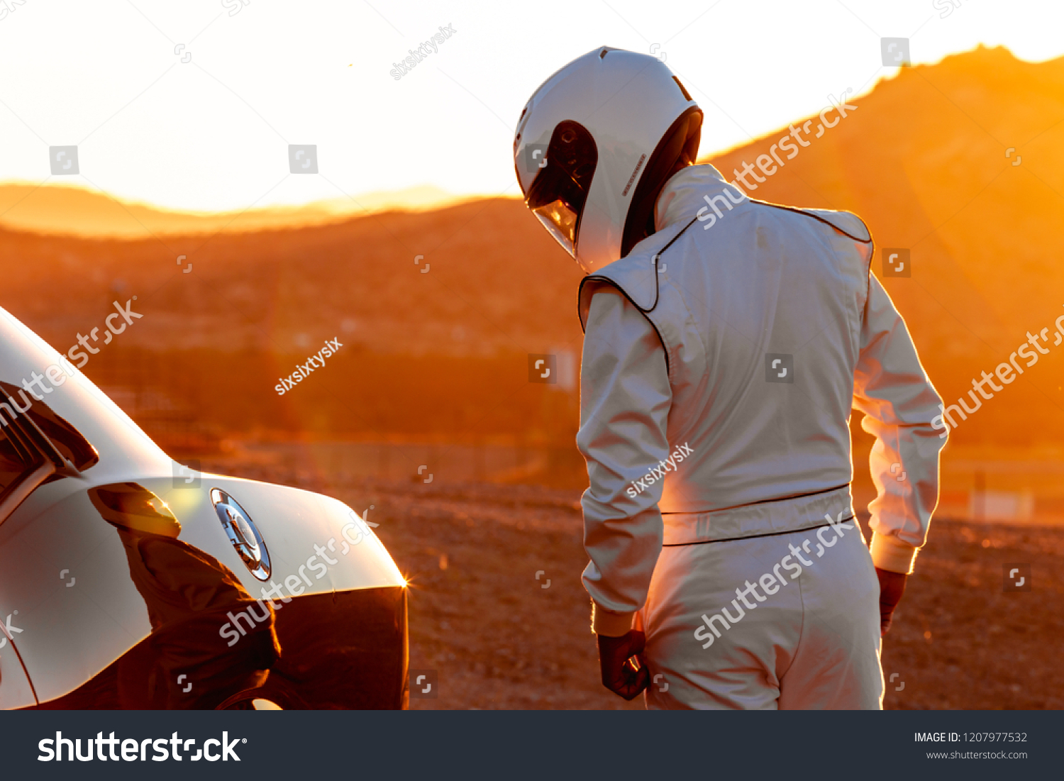 A Helmet Wearing Race Car Driver In The Early Morning Sun Looking At His Car Before Starting #1207977532