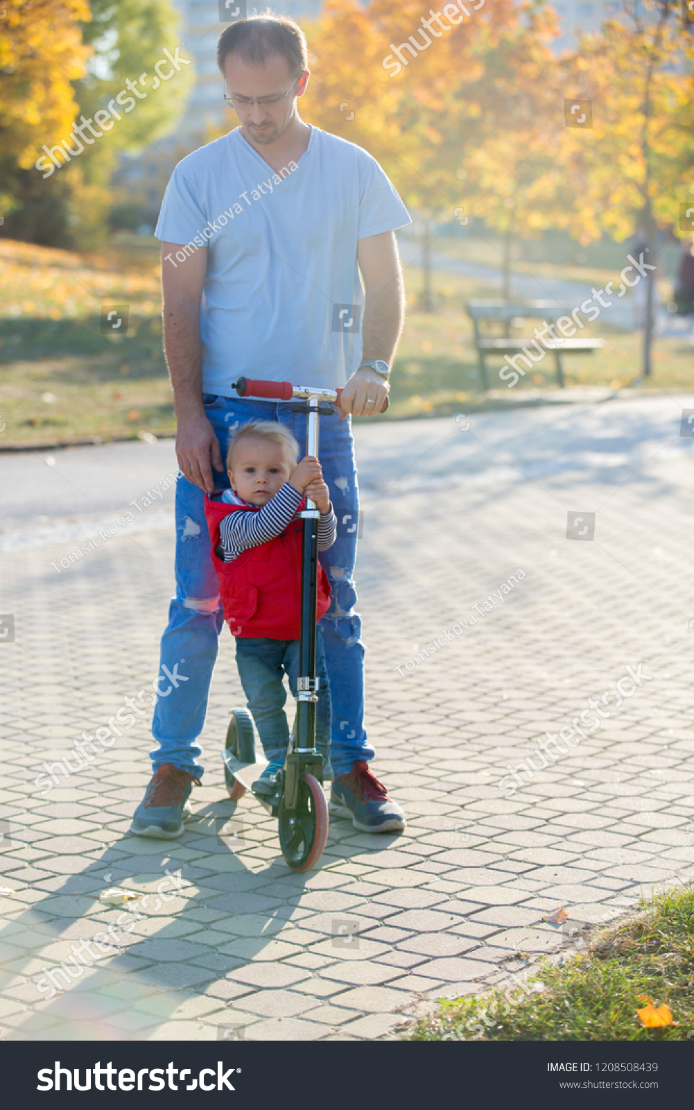 Father riding with his toddler son  scooter in a autumn park. . Active family leisure. Sports, leisure with kids concept. #1208508439