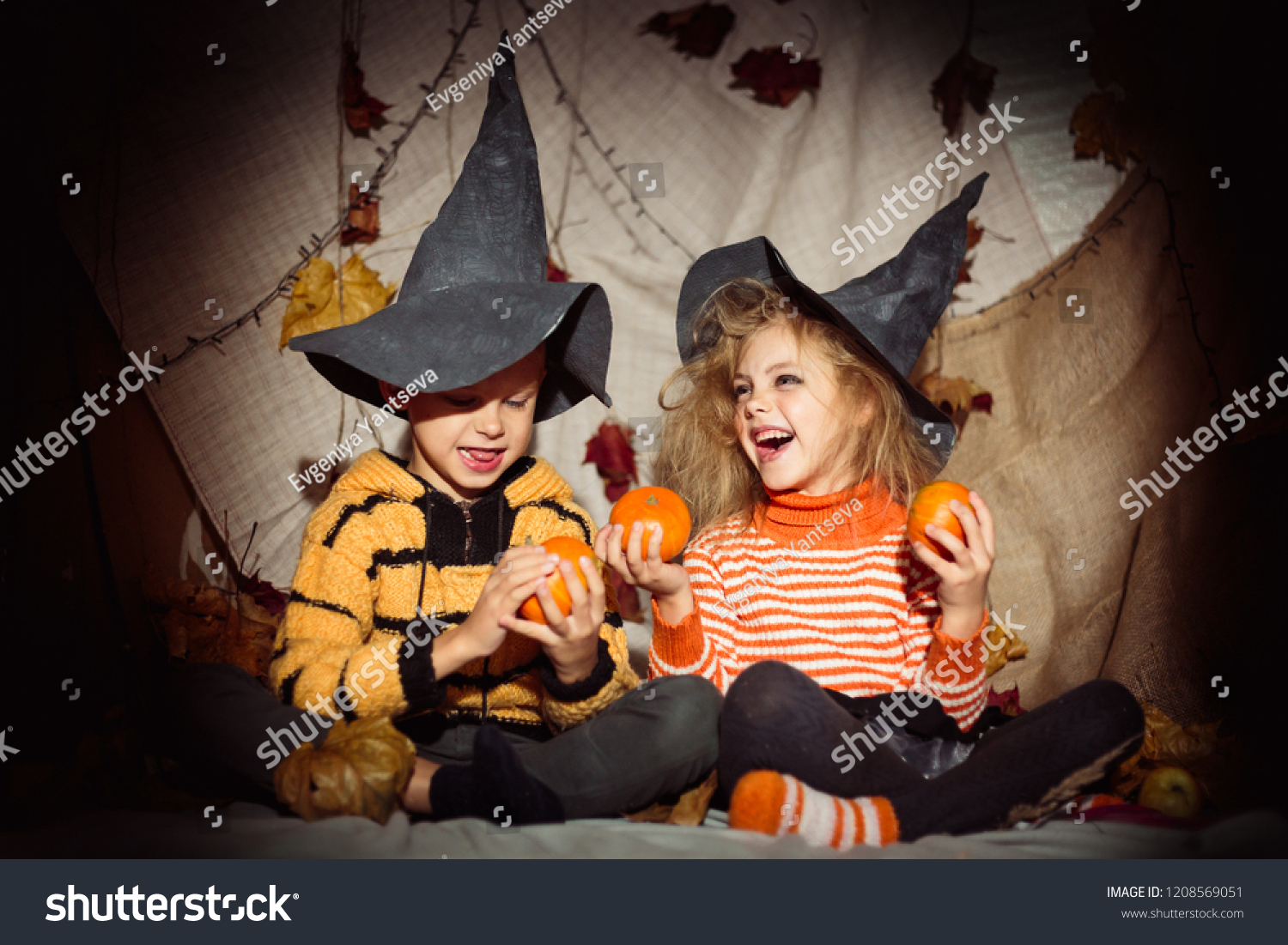 small happy children witches and witches in black hats and bright clothes halloween, boy and girl look at each other and hold pumpkins in hands and laugh #1208569051