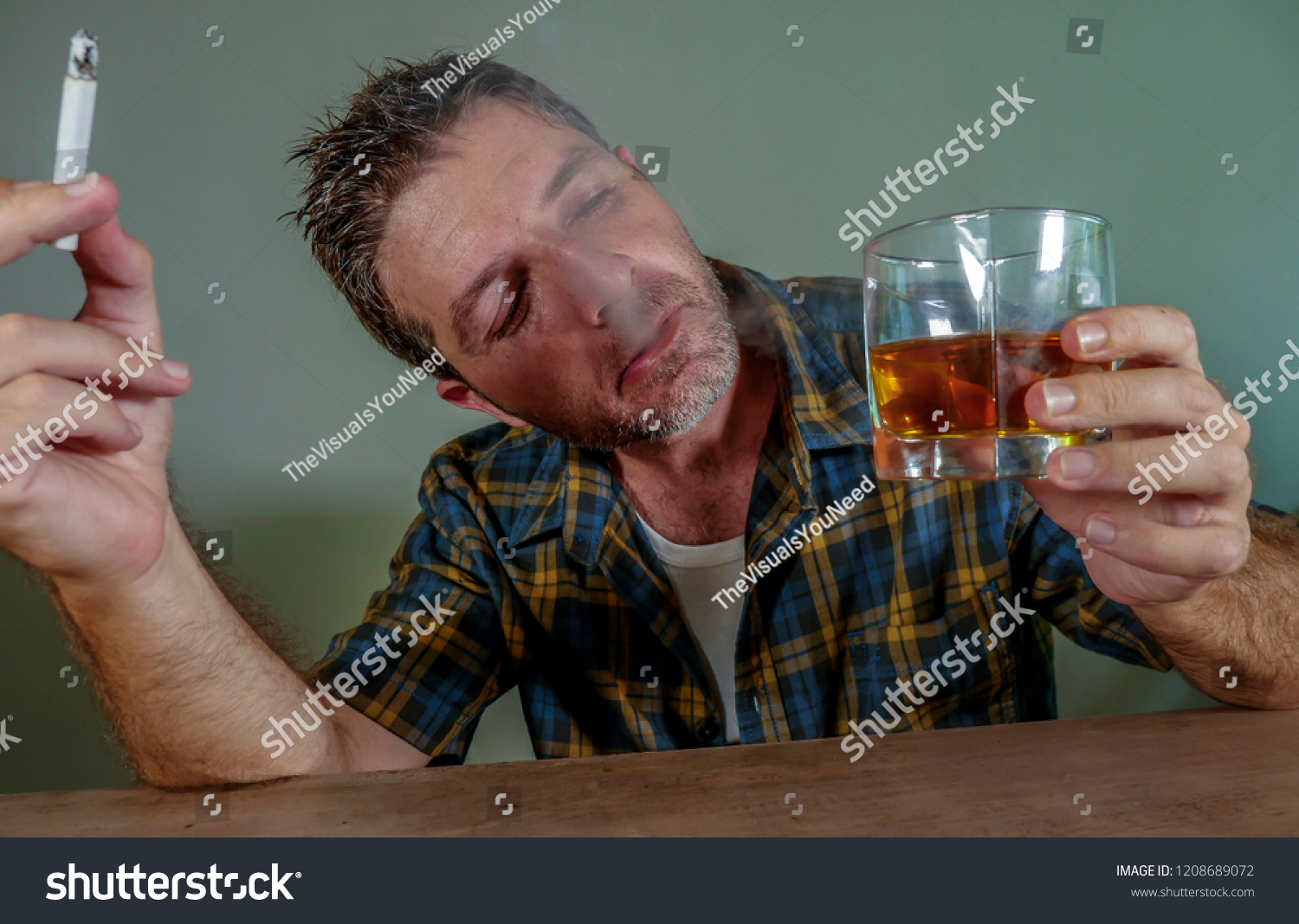 young messy and wasted addict man smoking cigarette having alcoholic drink looking at whiskey glass suffering alcoholism and tobacco addiction in unhealthy habit concept and alcohol abuse #1208689072