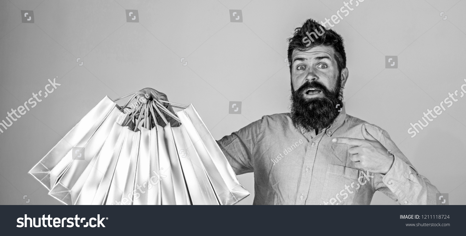 Boyfriend carrying lots of shopping bags and pointing at bags. Man with beard carries bunch of shopping bags, grey background. Recommendation concept. Hipster on surprised face recommends to buy. #1211118724