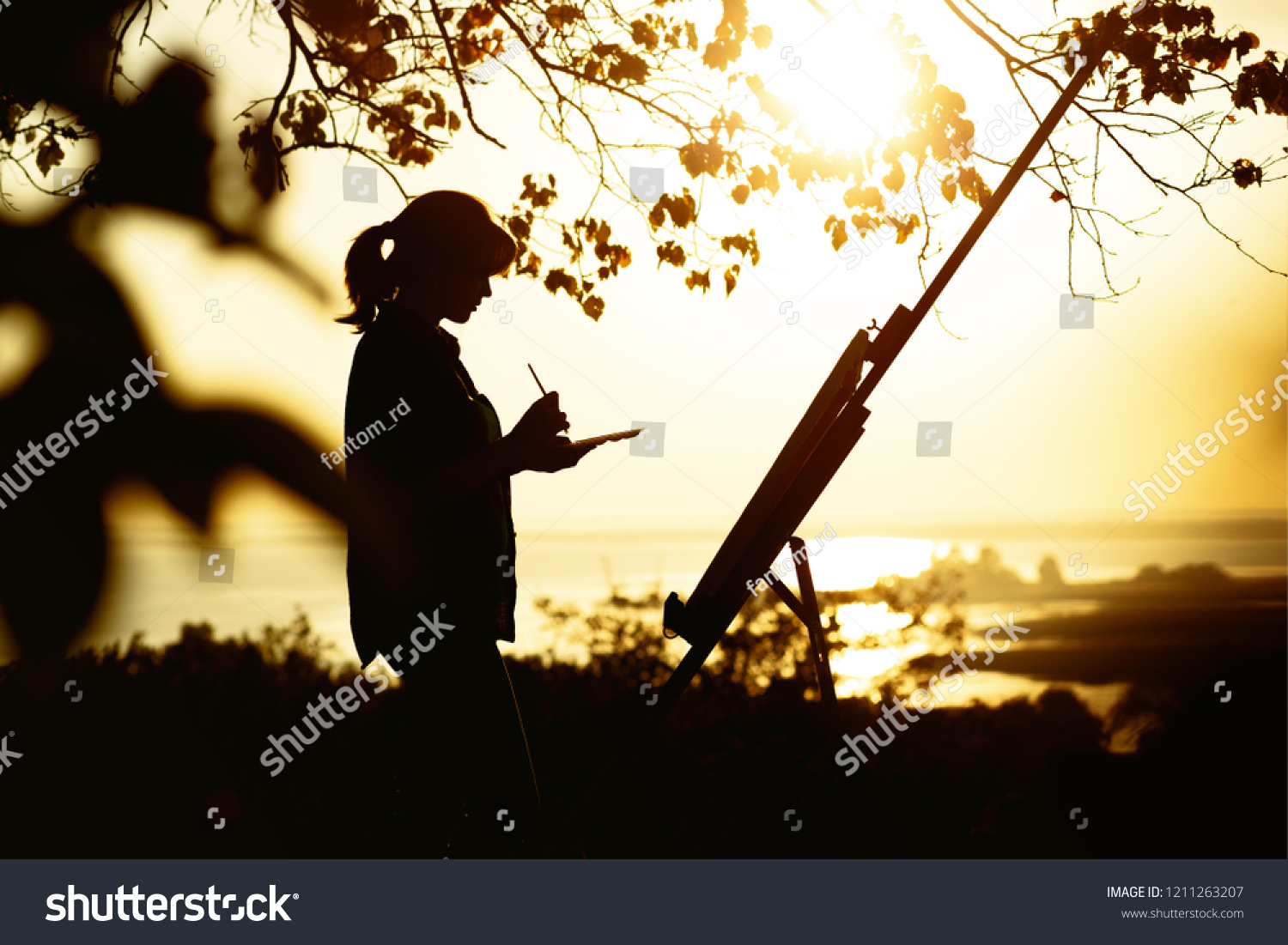 silhouette of a young woman painting a picture with paints on canvas on an easel outdoors, girl profile with paint brush and palette engaged in art on the nature in a field at sunset #1211263207