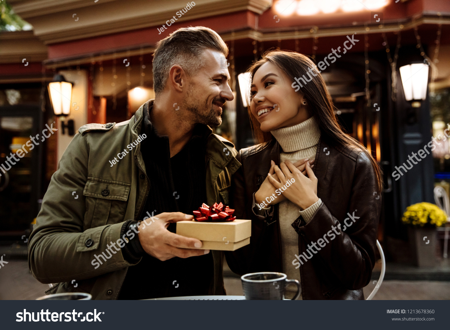 Couple. Holiday. Cafe. Man is giving a gift box to his woman. Both are in warm casual clothes smiling while sitting in the cafe outdoors #1213678360