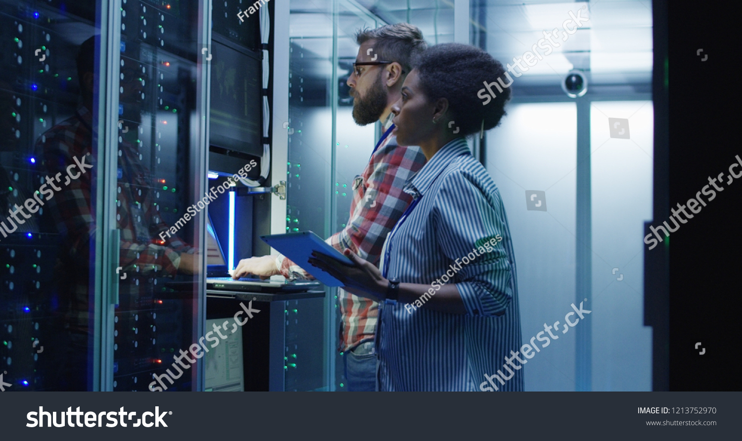 Modern multiethnic man and woman with tablet using laptop in server room while checking servers #1213752970